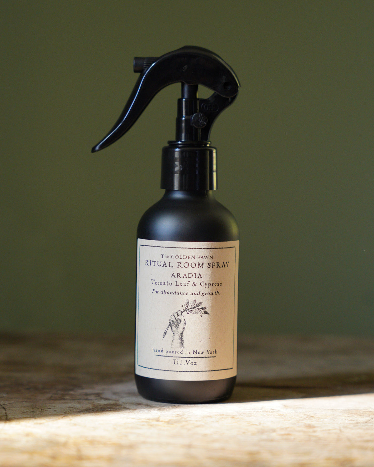 A dark amber spray bottle of Ritual Room Spray in the Aromat note, featuring a black trigger sprayer and a cream label with a zebra illustration, set on a rustic wooden surface against a muted olive green background.