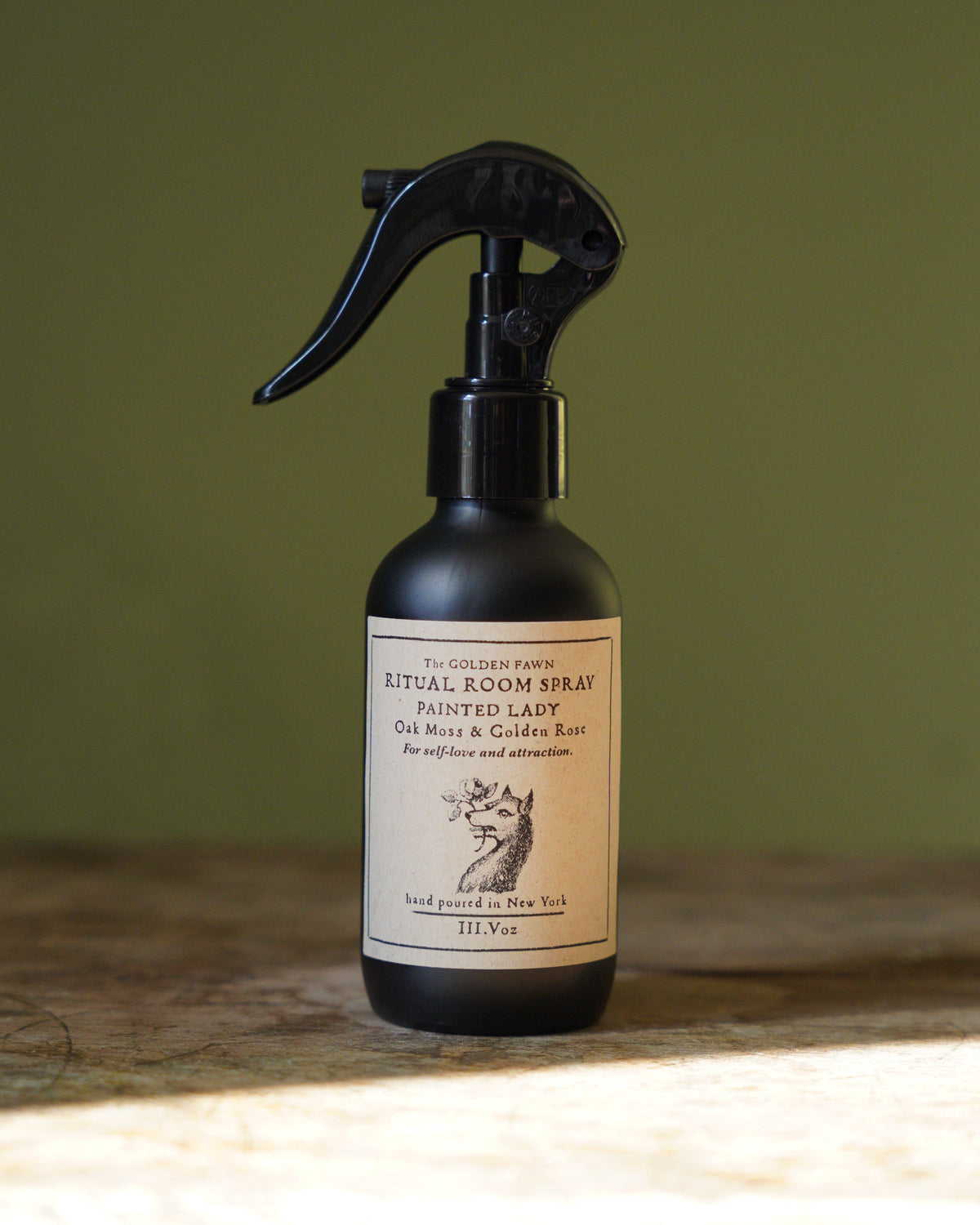 A dark amber glass spray bottle of Royal Room Spray with a black trigger sprayer and a cream-colored label, set on a rustic wooden surface against a muted olive green background.