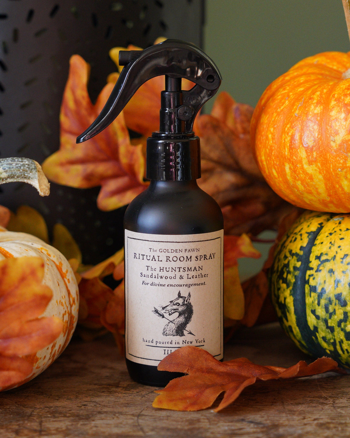 A bottle of Ritual Room Spray Conditioner stands on a rustic wooden table surrounded by decorative pumpkins, orange autumn leaves, and a dark metal lantern against a muted green wall.