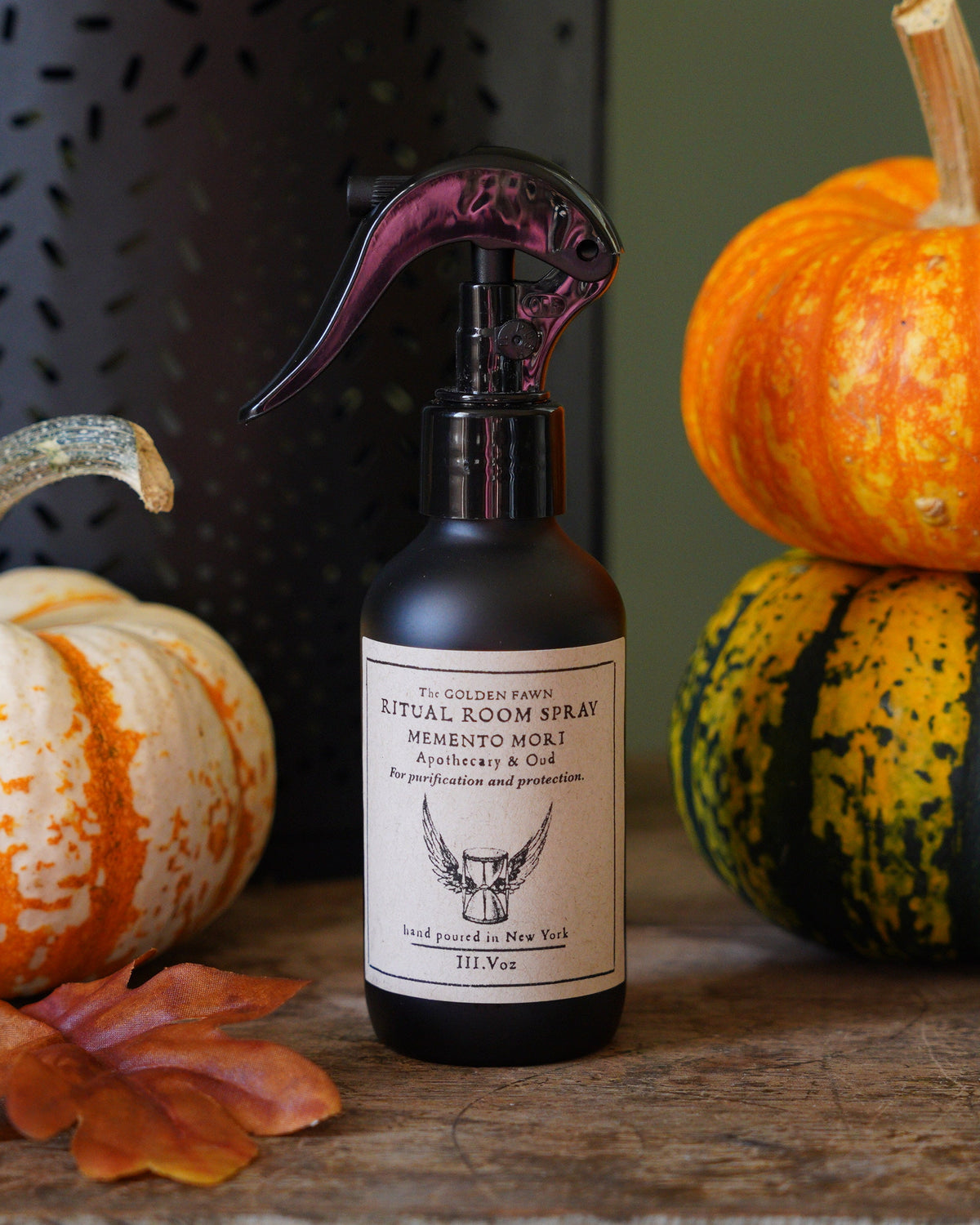 A bottle of Ritual Room Spray with a black sprayer and white label, styled on a rustic wooden table with decorative pumpkins, autumn leaves, and a dark metal lantern against a muted green background.