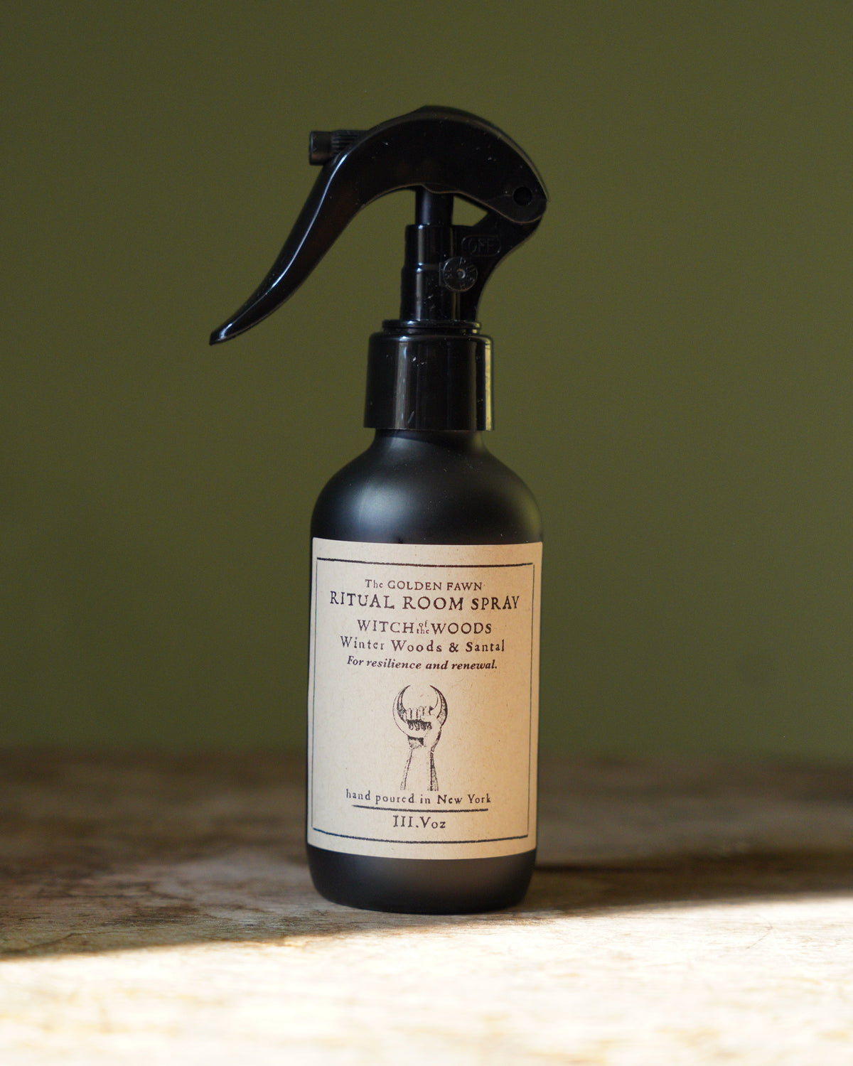 A 200ml spray bottle of The Artisan Room Spray with a black trigger sprayer and cream-colored label, set on a rustic wooden surface against a dark olive green background.